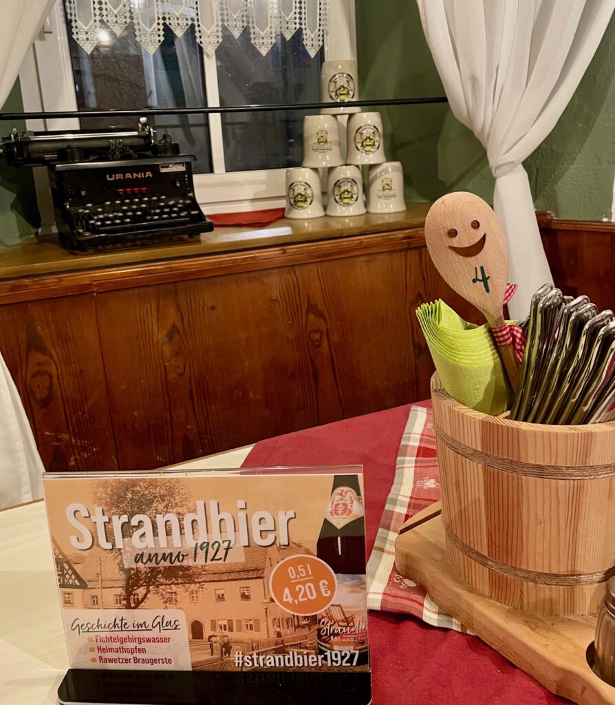 Gaststumm-Aufnahme aus der Brauereigaststätte am Strand mit Strandbier-Hinweisschild, Besteckfass sowie alten Bierkrügen und einer historischen Schreibmaschine auf dem Fensterbrett.